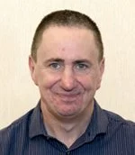 Portrait of a man with short light brown hair, blue eyes, and a light skin tone, smiling, wearing a dark collared shirt.