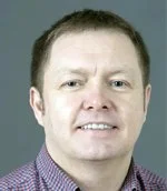 A headshot of a man with short reddish hair, fair complexion, wearing a collared shirt, smiling at the camera.