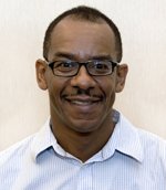 Portrait of a man with glasses, smiling, wearing a light blue dress shirt.