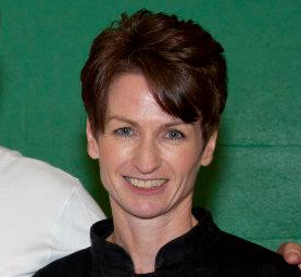 A woman with short brown hair smiling, wearing a black top, against a green background.