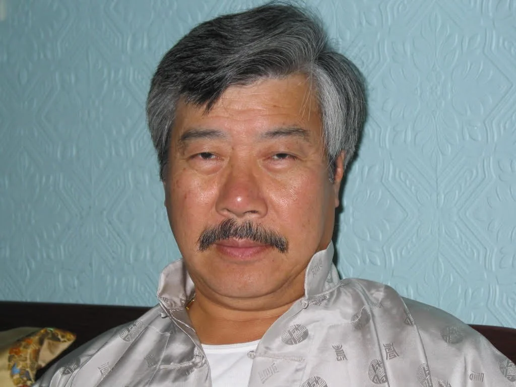 Close-up of an older man with gray hair and a mustache, wearing a light-colored satin shirt, sitting against a textured light bluish wall.