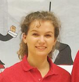 A young woman with curly hair, smiling, wearing a red shirt, standing in front of a colorful mural.