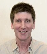 A smiling man with short dark hair wearing a light-colored shirt