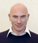Bald man wearing a white shirt and dark sweater, smiling slightly, indoor setting with light-colored background.