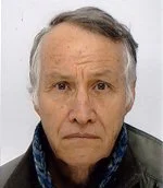 Close-up of an elderly man with gray hair, light skin, and a serious expression, wearing a dark jacket and a blue shirt, against a plain white background.