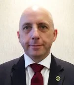 A bald man in a black suit, white shirt, and red tie posing against a plain background.