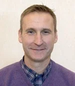 A man with short light brown hair, wearing a purple sweater over a checkered shirt, smiling and looking at the camera. Plain beige wall background.
