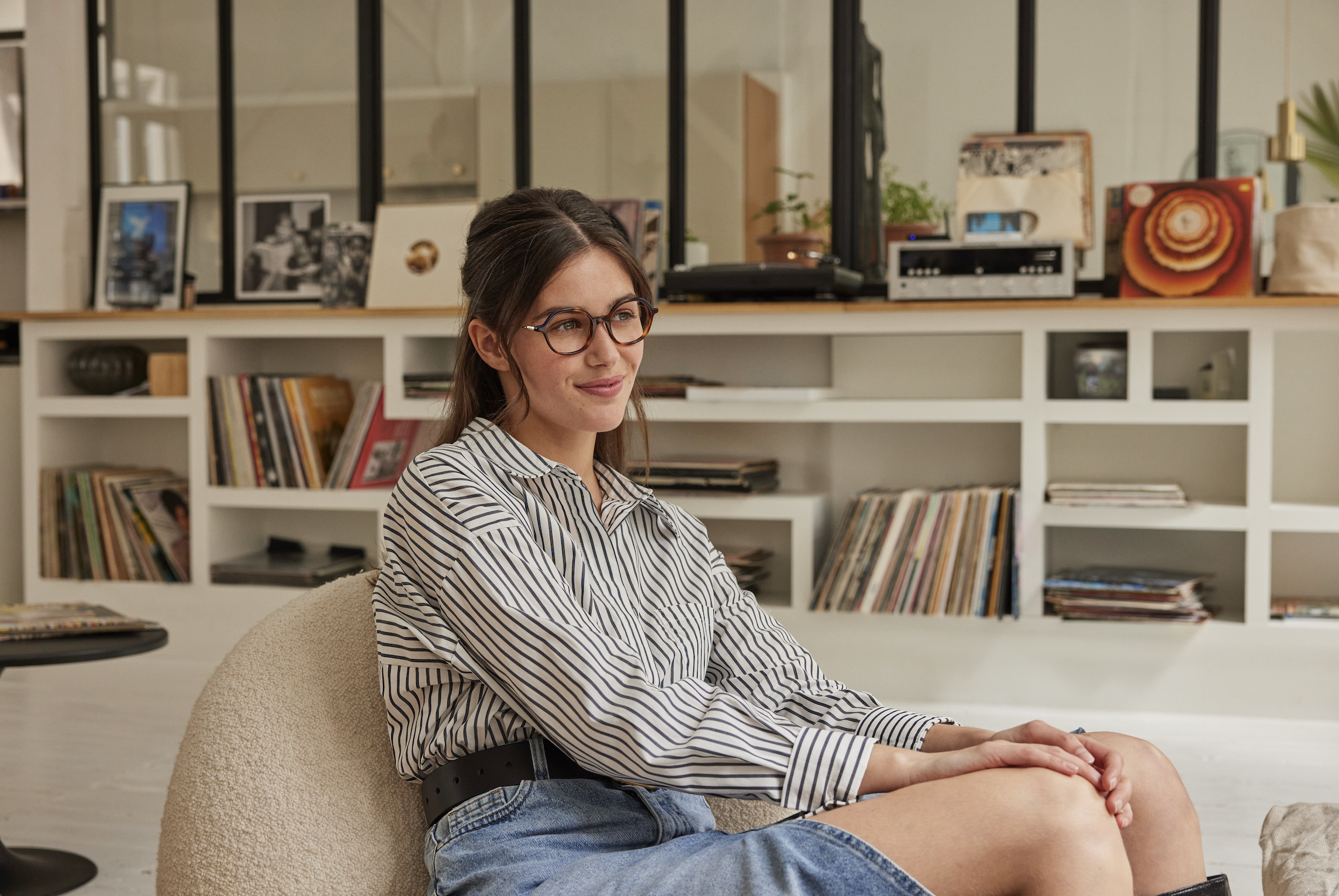 Een jonge vrouw met bril en lang haar zit in een kamer, gekleed in een gestreept overhemd en een denim rok, met een achtergrond van boekenplanken, platen en kunstwerken.