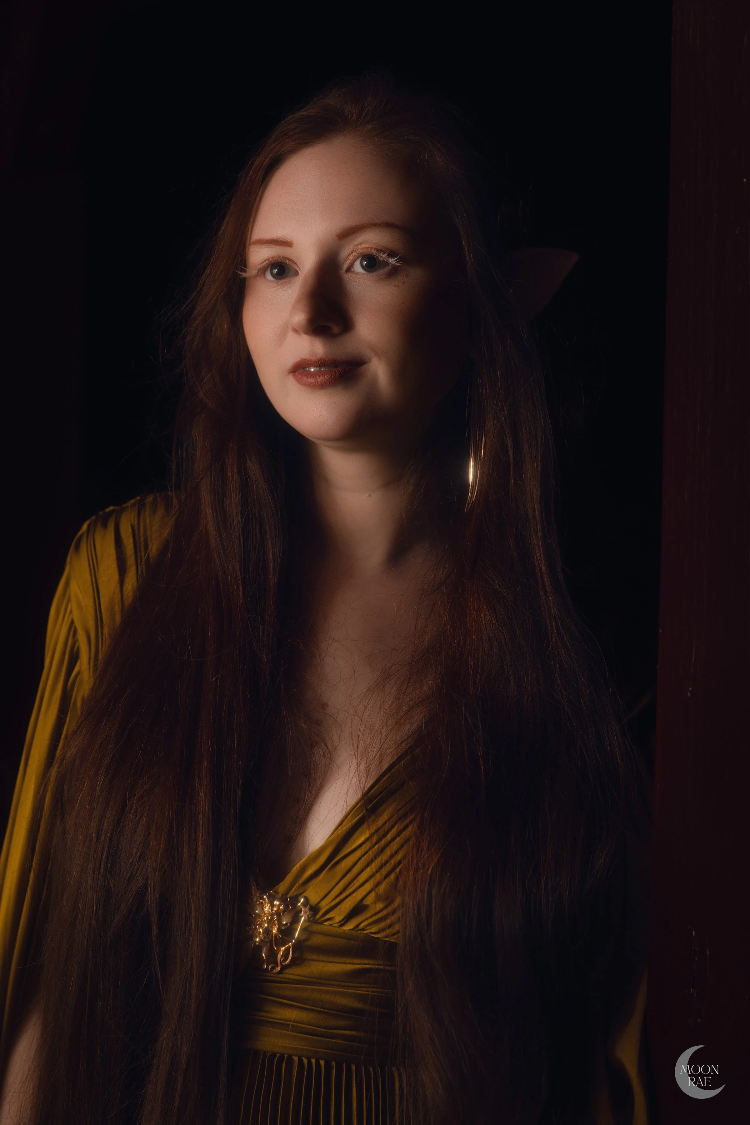 A woman with long auburn hair and blue eyes, wearing a gold dress with a brooch, standing near a wooden surface in low lighting.