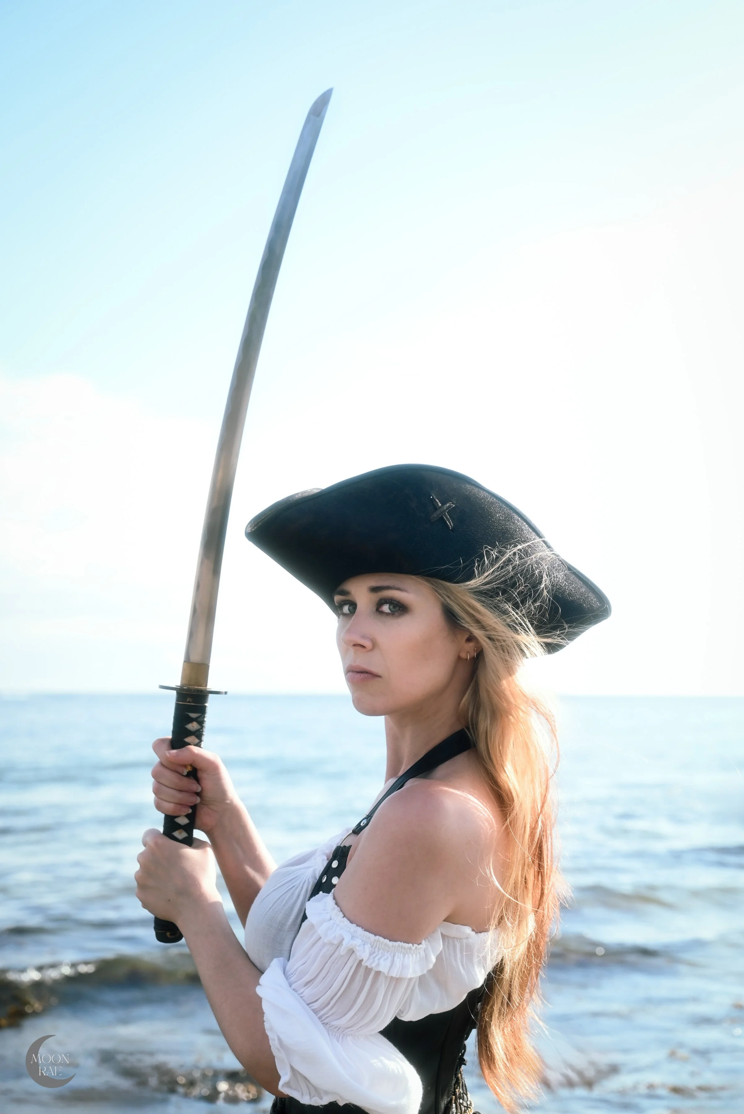 A woman dressed as a pirate with a black tricorn hat holding a sword stands on the beach with the ocean in the background.