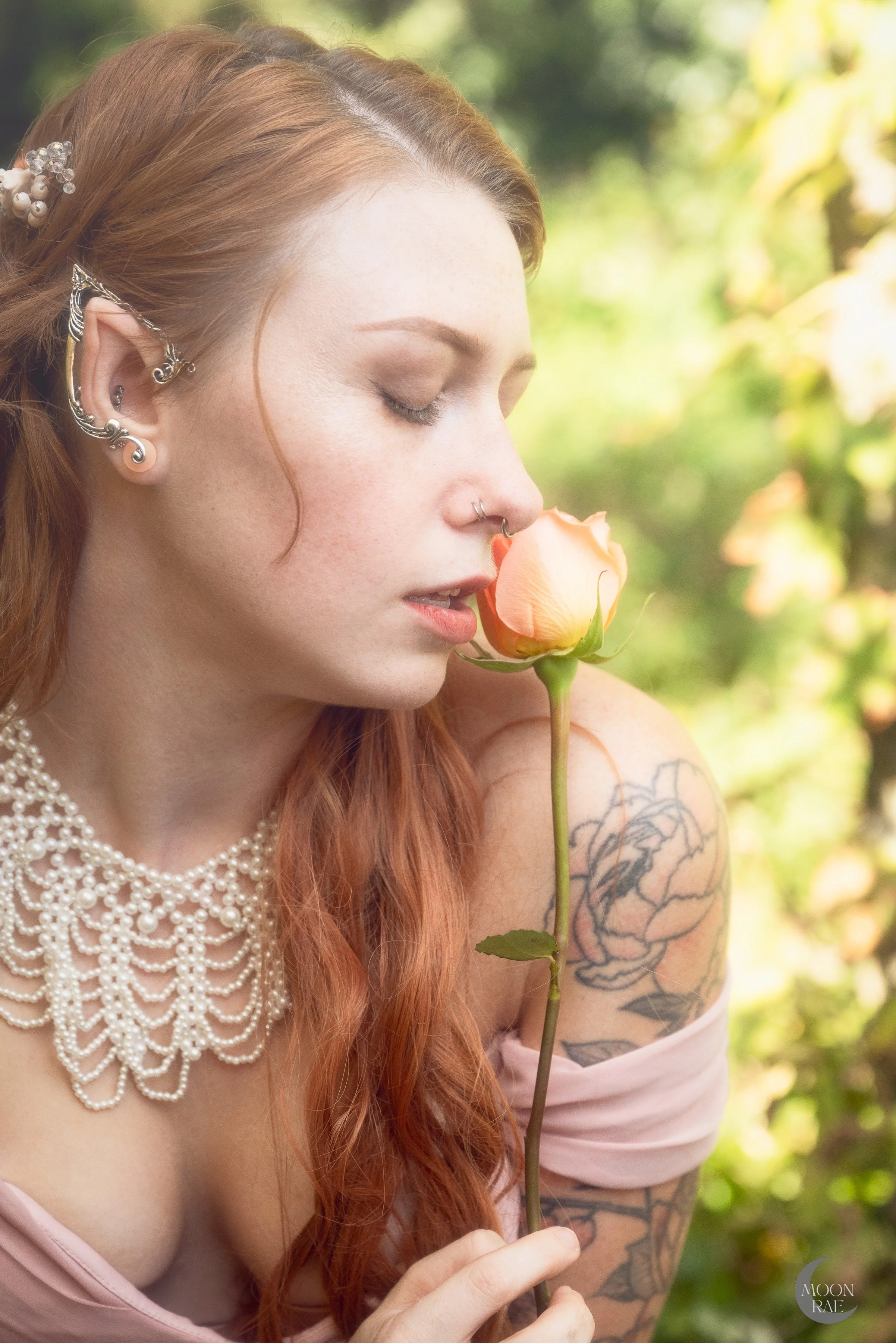 A woman with red hair and multiple earrings holds a peach-colored rose close to her face, with her eyes closed outdoors with blurred green foliage in the background.