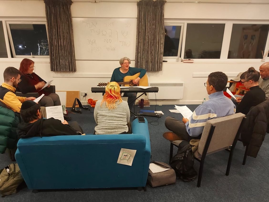 Mich is playing guitar in a classroom, surrounded by students holding papers.