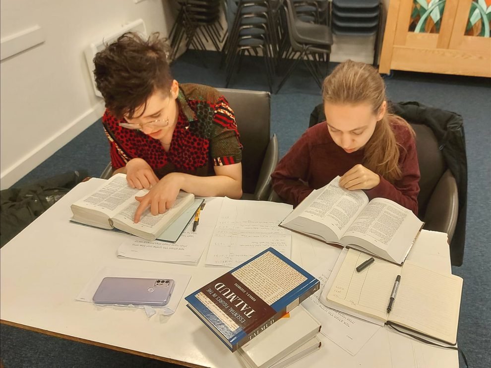 Jo and another student are sat around a table in a classroom, finding a word in their dictionaries. There is a book that says TALMUD on the table, as well as notebooks. They are studying intently!