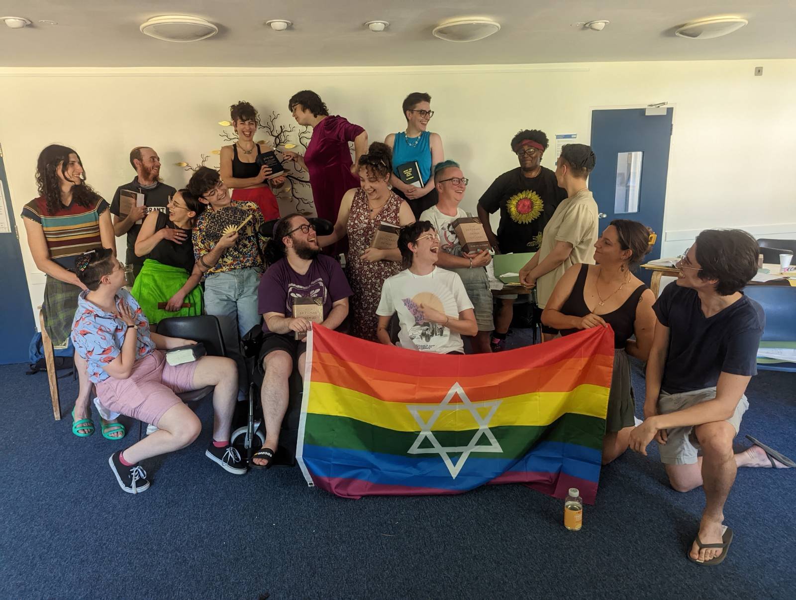 A bunch of queer Jews are stood around a pride flag with the Star of David in the middle, posing for the photo. However, they are mainly laughing and happy.