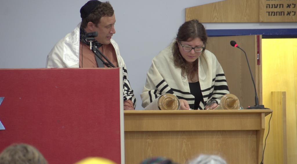 Rabbi Lev stands ready to help a Bnei Mitzvah student read from Torah. They are both wearing tallitot striped with blue.
