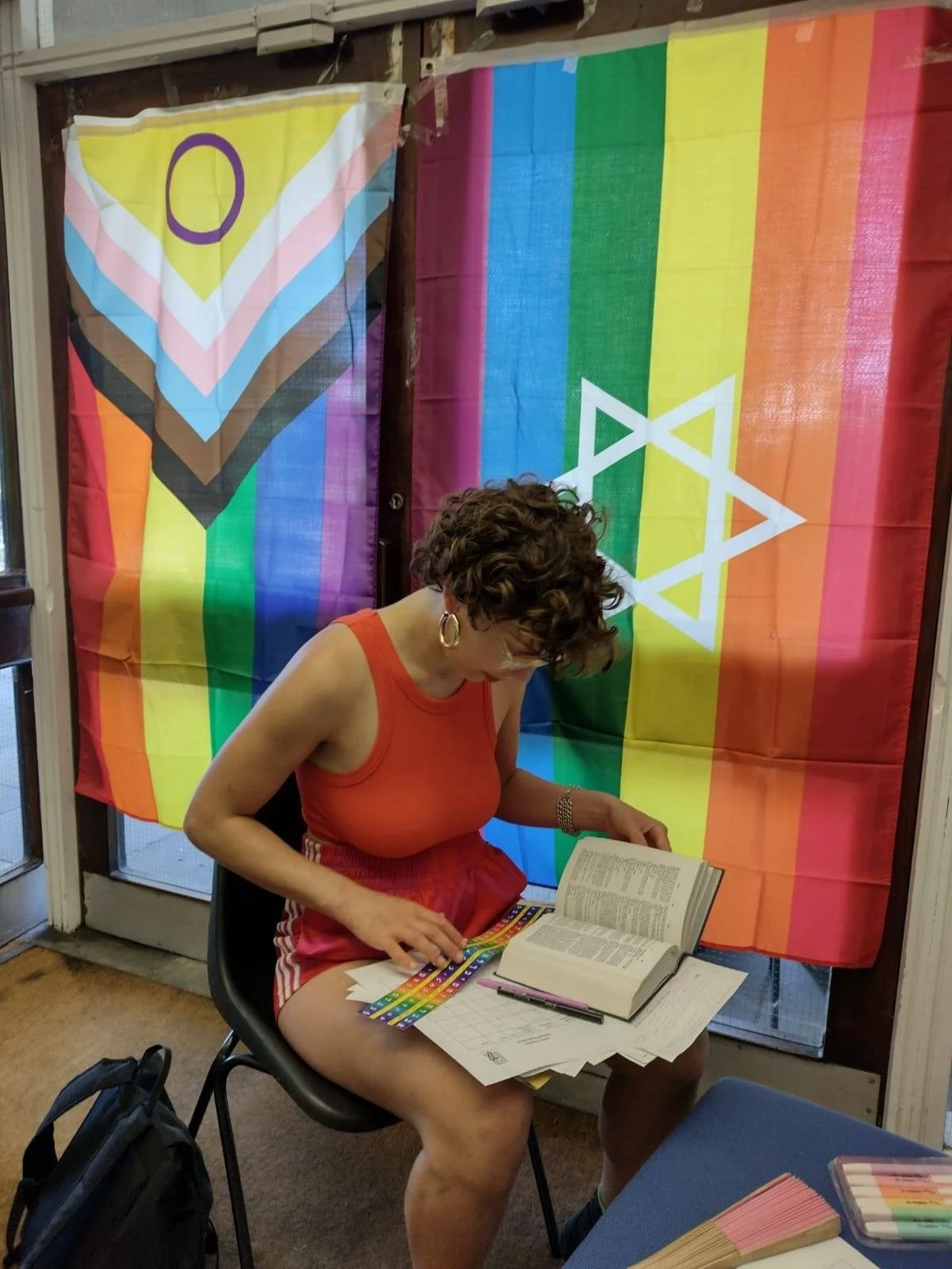 A white Jewish person is sat at a chair studying, with a dictionary open. They are holding a pen. Behind them is two rainbow flags, one with the intersex, trans, and Black and brown colours and one with a Star of David on it.