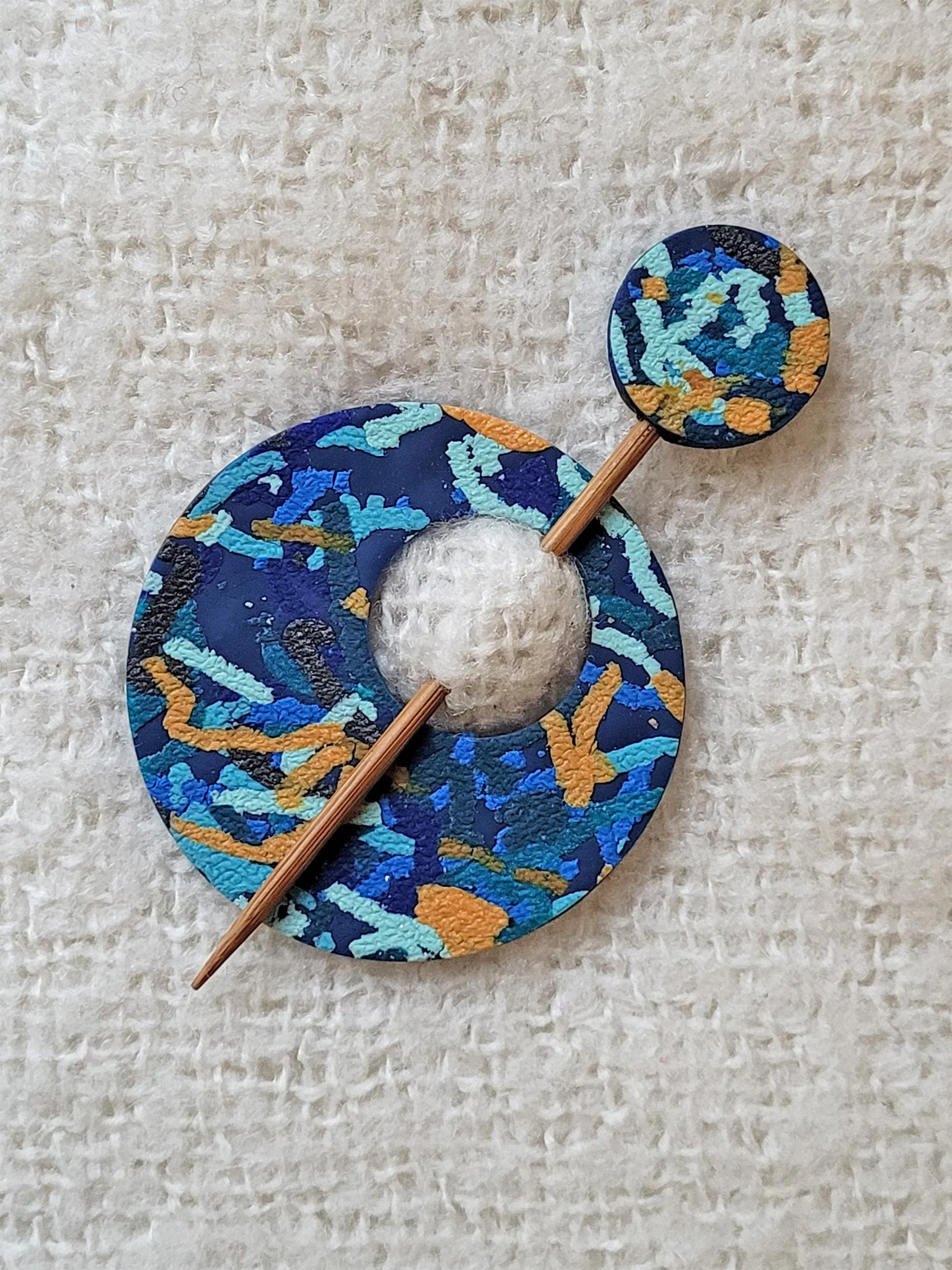 Flat lay image of circular Blue Ticker Tape polymer clay shawl pin, with a bamboo pin, topped with a matching round disc.