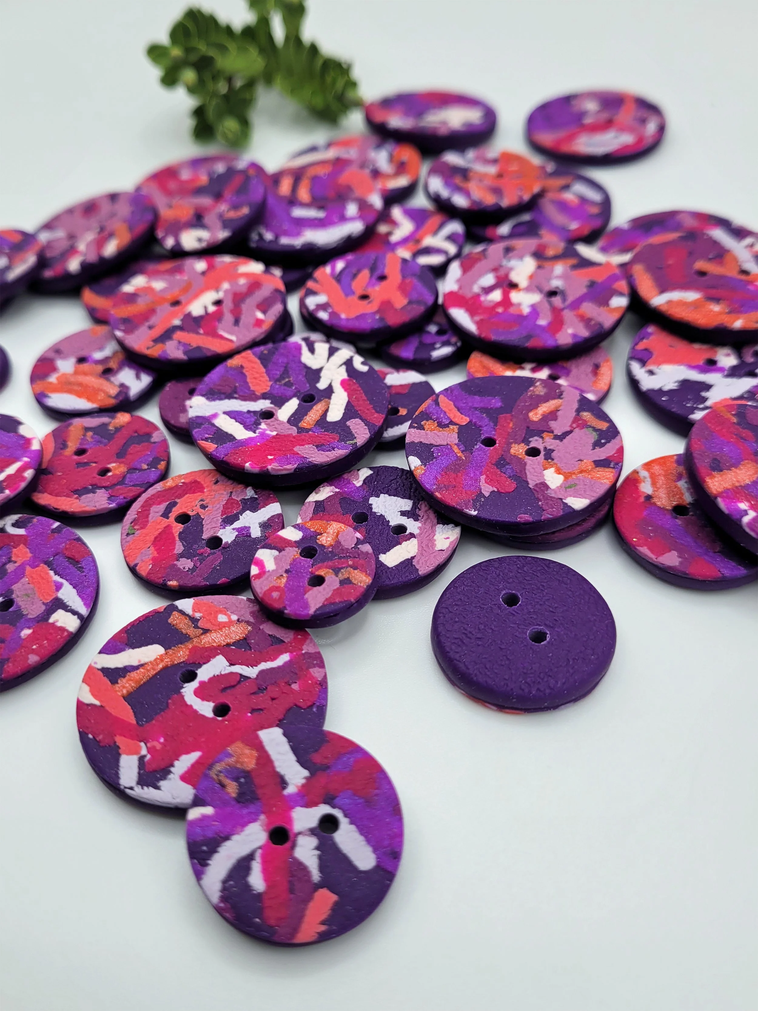 Purple Ticker Tape buttons scattered on a white surface with a small green plant the background.