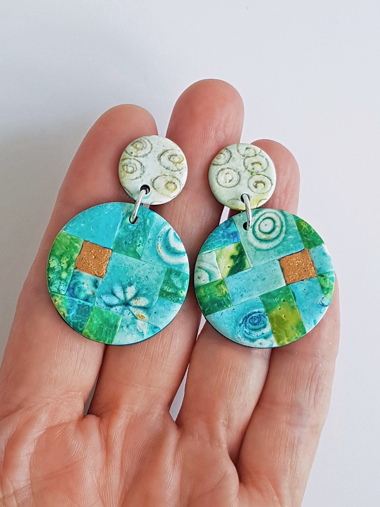 Faux bronze leaf accent tiles in turquoise, blue mosaic disc beads, hanging from cream and gold stud tops, held between fingers.