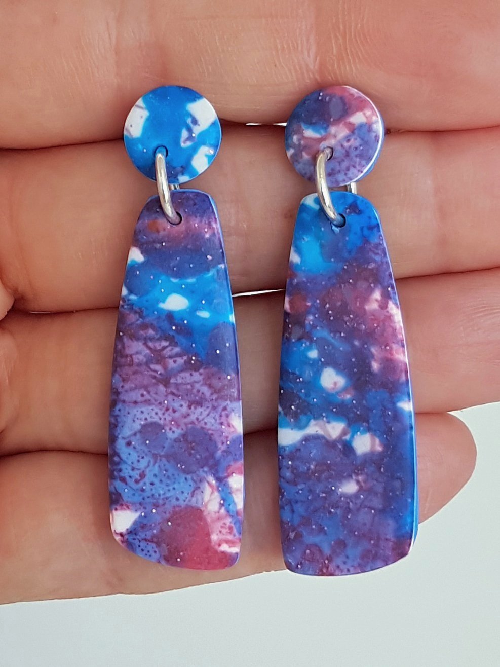 Blue, red and white mismatched dangle earrings with a matte, line and brushstroke pattern held between fingers.