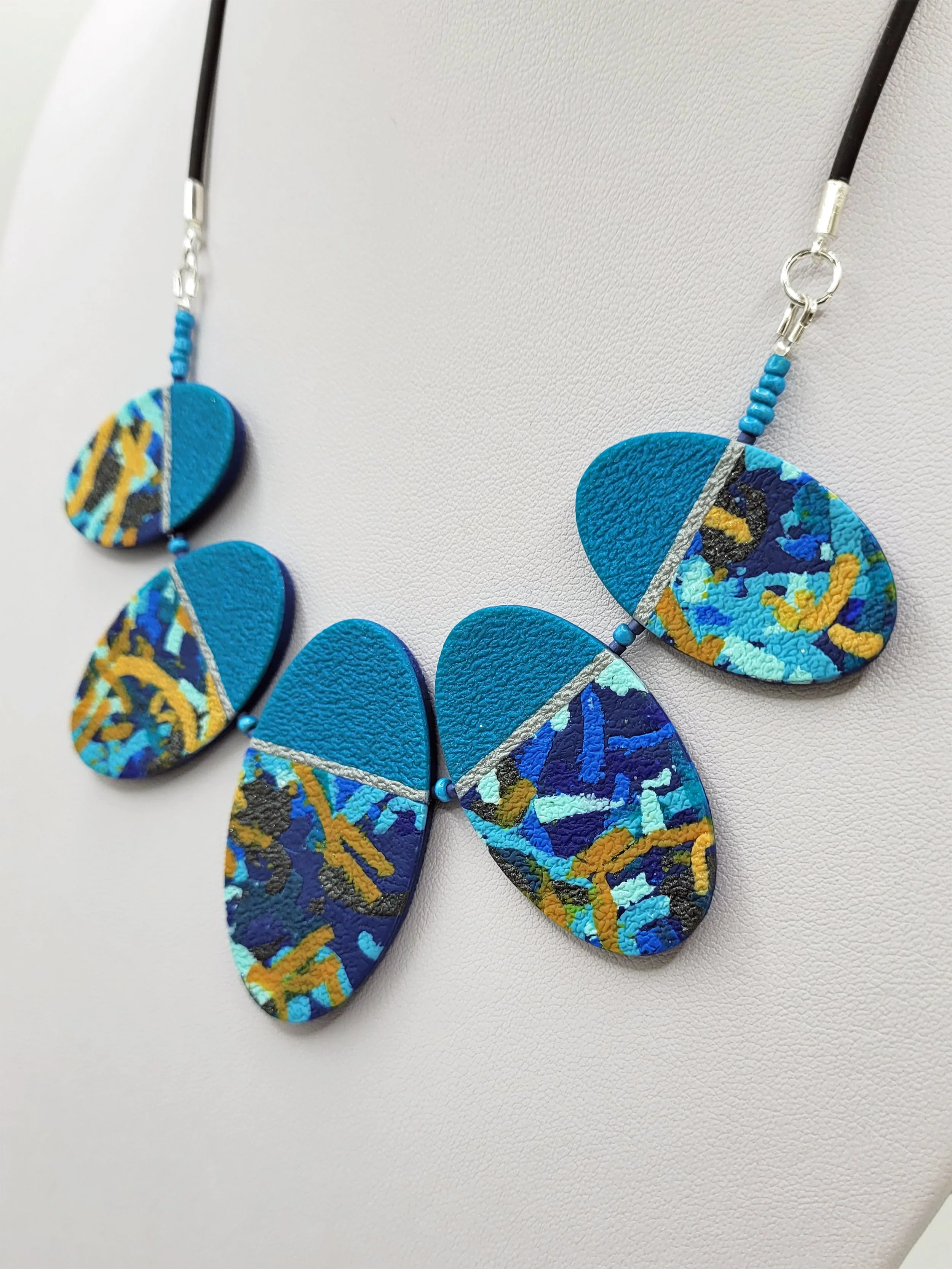 Each oval bead features a pearlescent peacock blue upper third separated by a thin metallic silver band from the lower two thirds of abstract ticker tape style pattern.