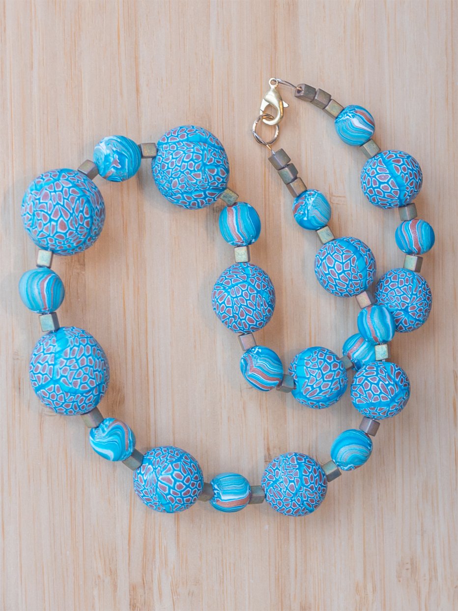 A flat lay image of a necklace with large and small handmade round beads decorated with a millefiori cane in turquoise, gold and white.