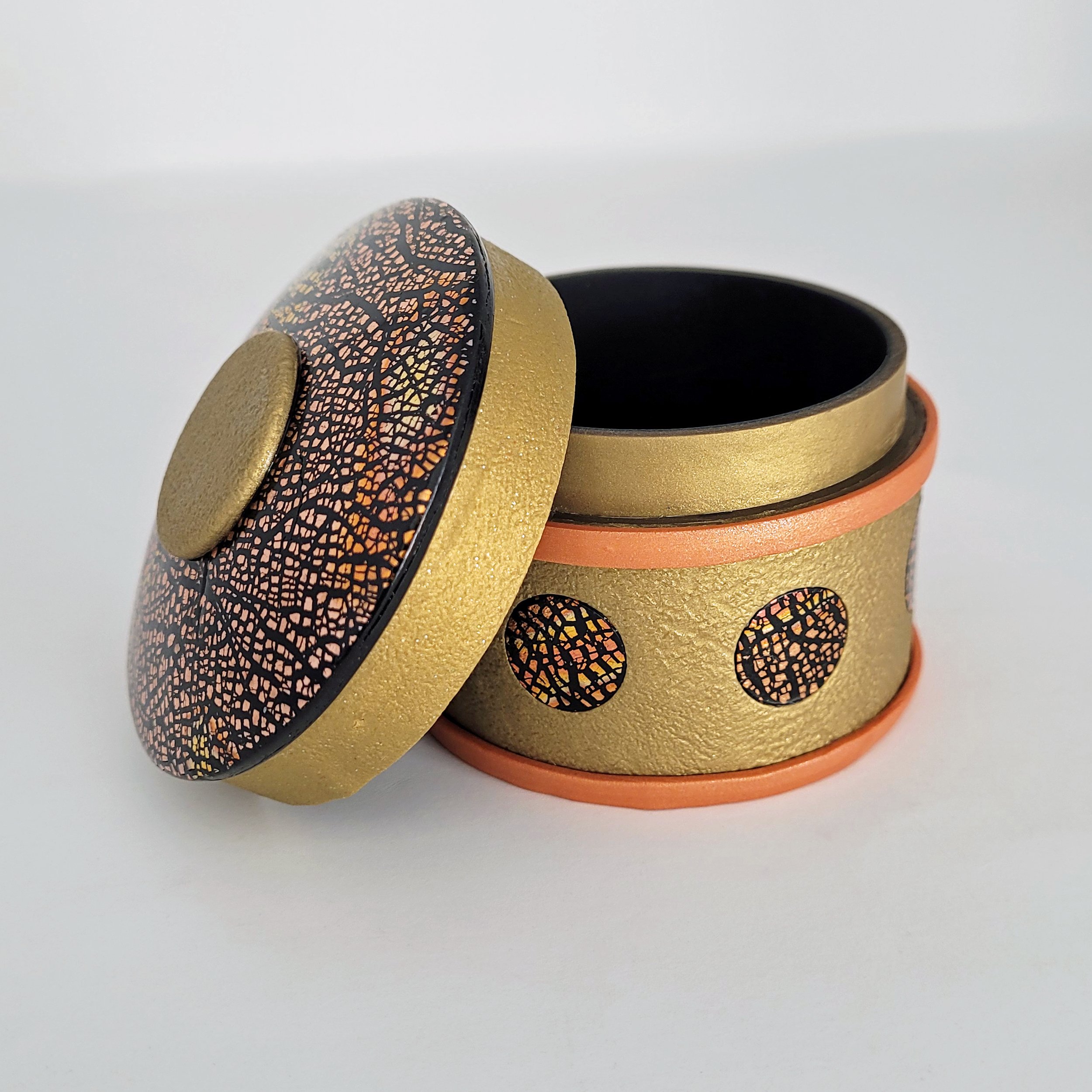 A shallow round box, in gold with a black lining, decorated with areas of crackled bronze, copper and black.