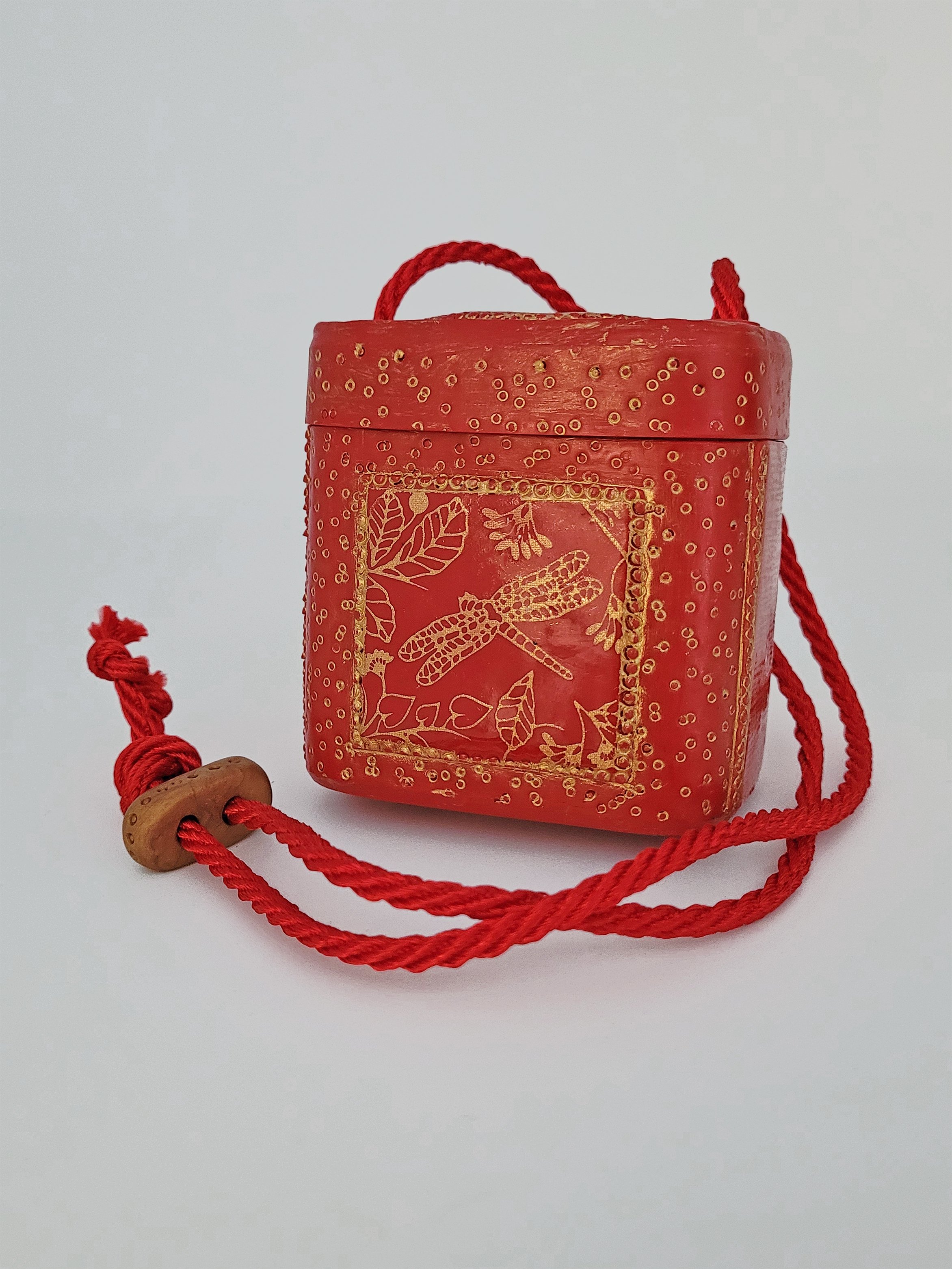 A red rectangular lidded box decorated with a gold dragonfly which can be carried with a red cord.