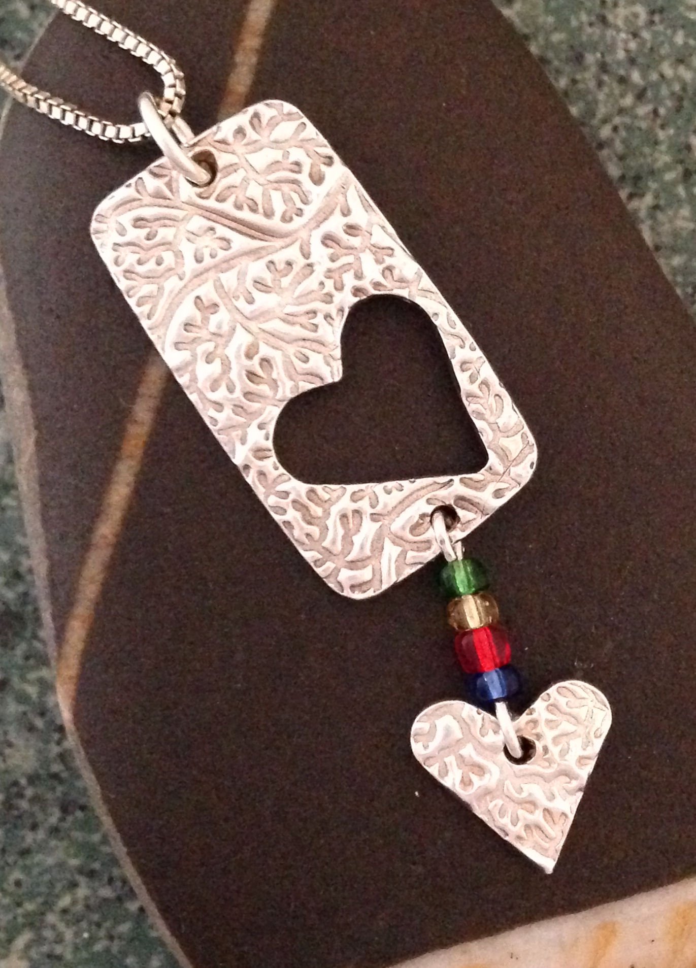 Embossed rectangular pendant with a heart cutout. The heart bead hangs from the bottom of the pendant.
