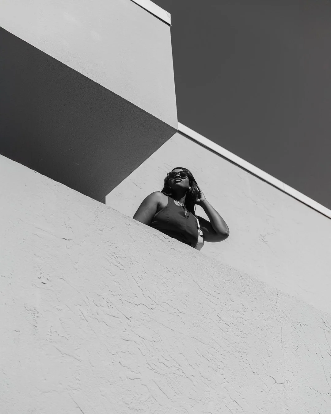 A woman wearing sunglasses and a sleeveless top standing on a balcony or ledge against a minimalist building with geometric lines, captured from a low angle in black and white.