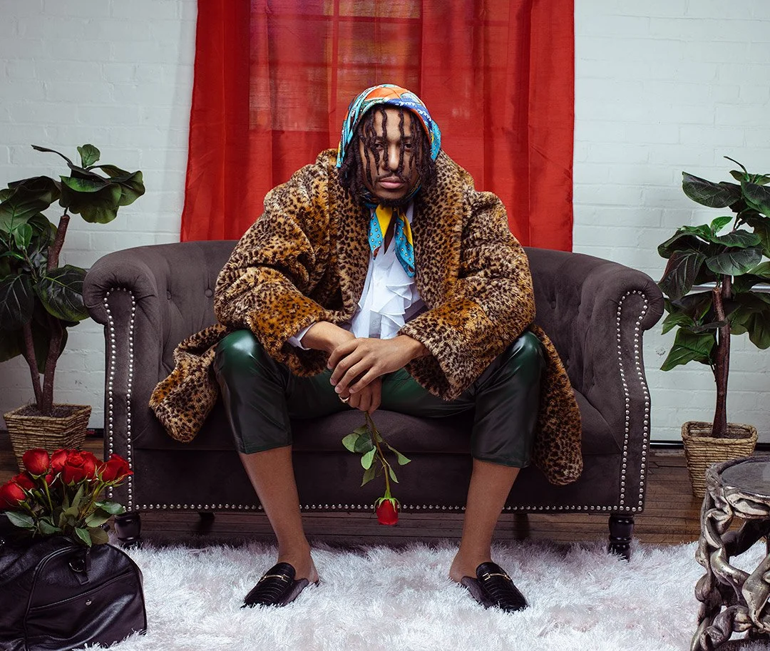 A person sitting on a dark sofa in a room with red curtains, holding a single red rose, wearing a leopard print coat, black pants, loafers, and a colorful headscarf, with plants and flowers around them.