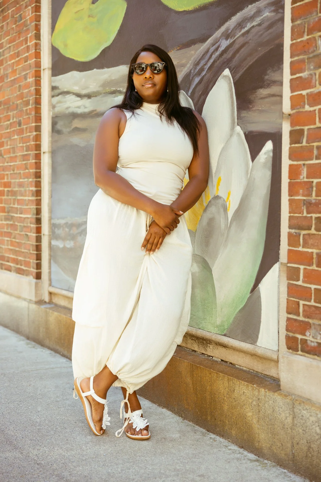 A woman posing outdoors against a painted wall of flowers and clouds, wearing sunglasses, a white sleeveless top, wide beige pants, and white shoes with bows.