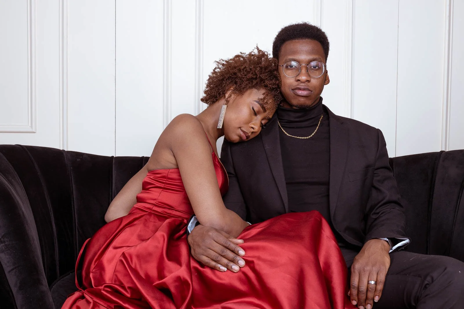A man and woman sitting on a black velvet sofa, with the woman resting her head on the man's shoulder, both dressed in formal attire. The woman wears a red dress and the man is in black with glasses and jewelry.