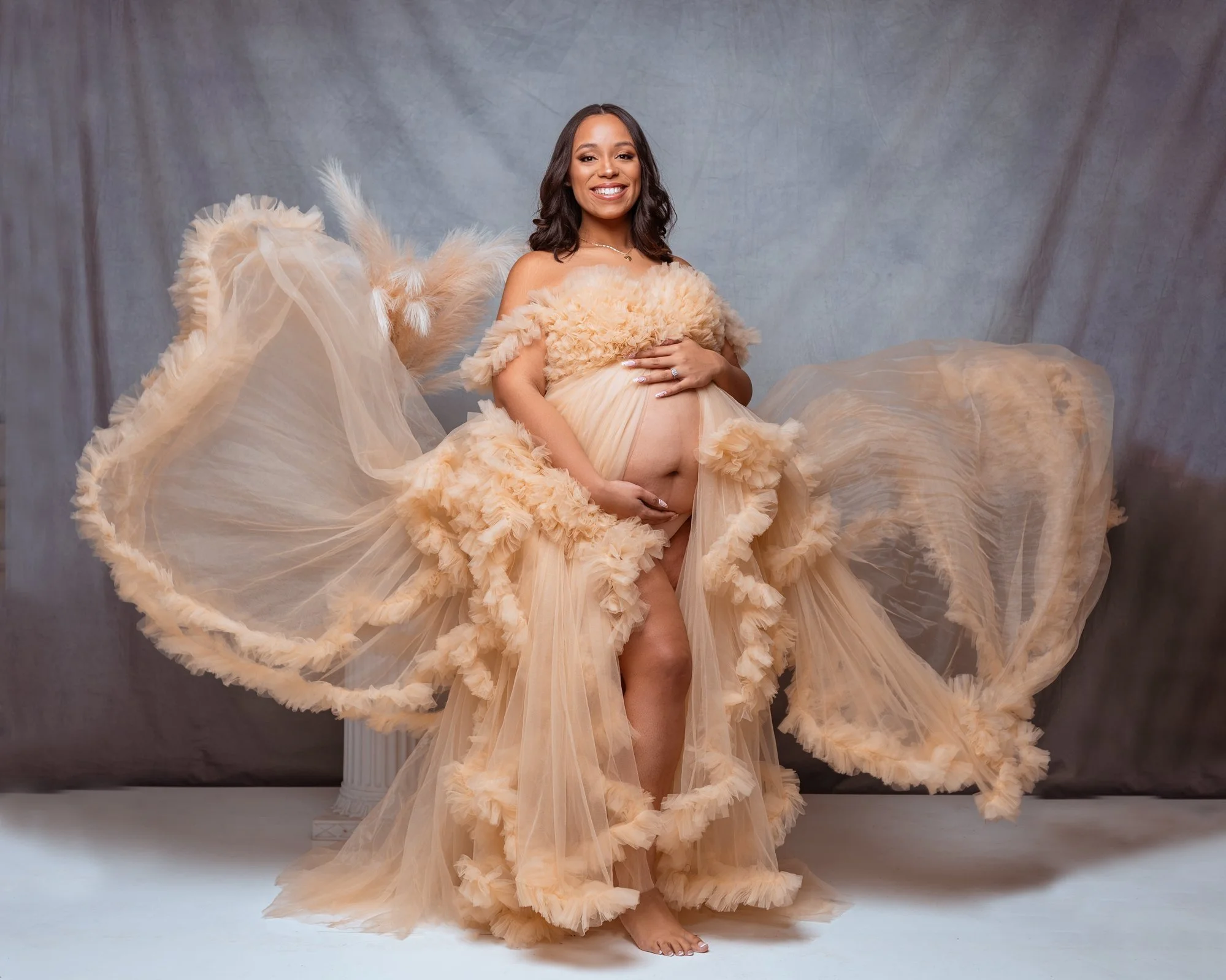 A pregnant woman with a happy smile, wearing an elegant peach-colored ruffled dress, holding her belly with one hand and a hand on her belly, standing against a gray backdrop.