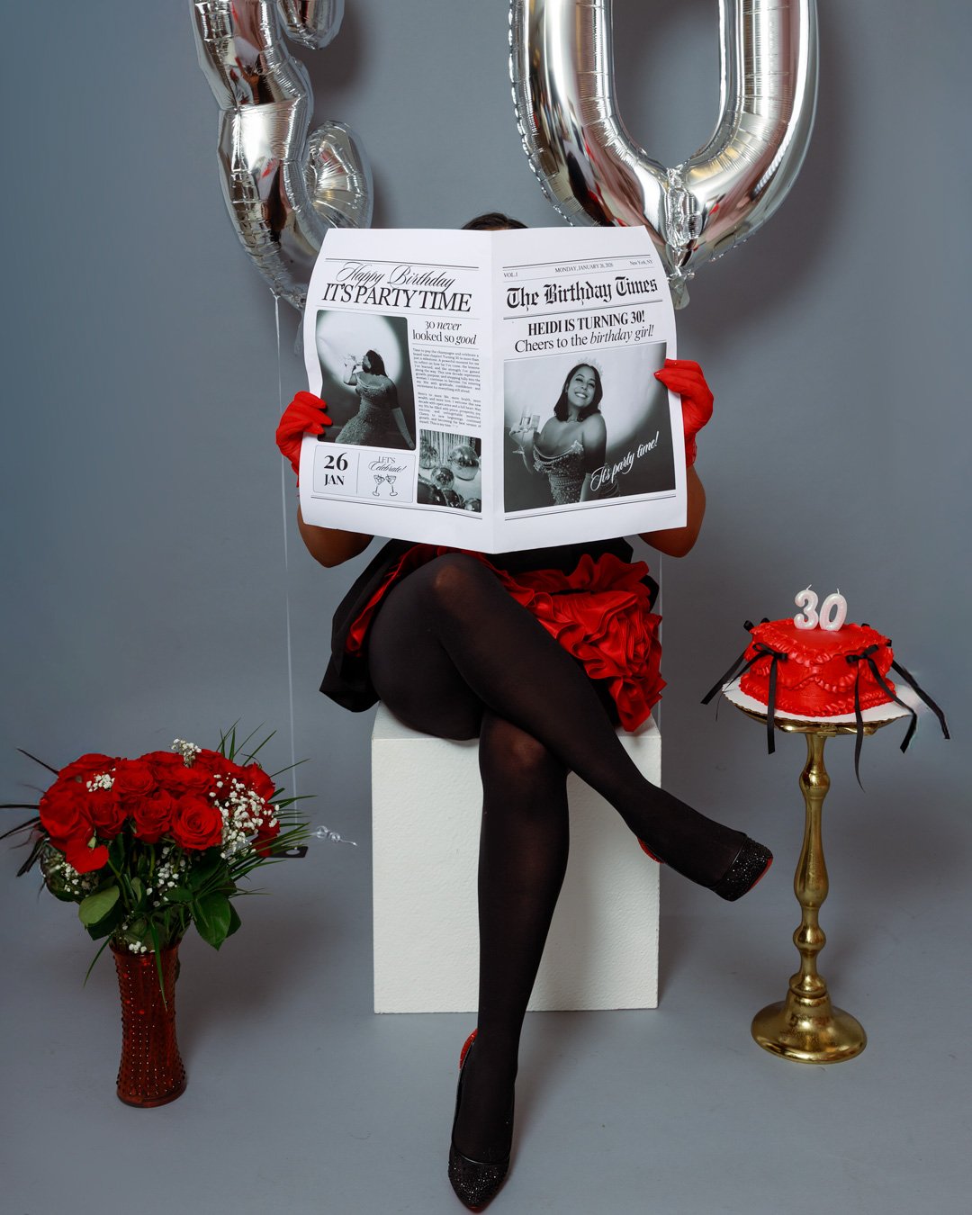 Person sitting on a white block, holding a birthday newspaper, wearing a red dress, black stockings, and red gloves, with balloons, a birthday cake, and floral bouquet around.
