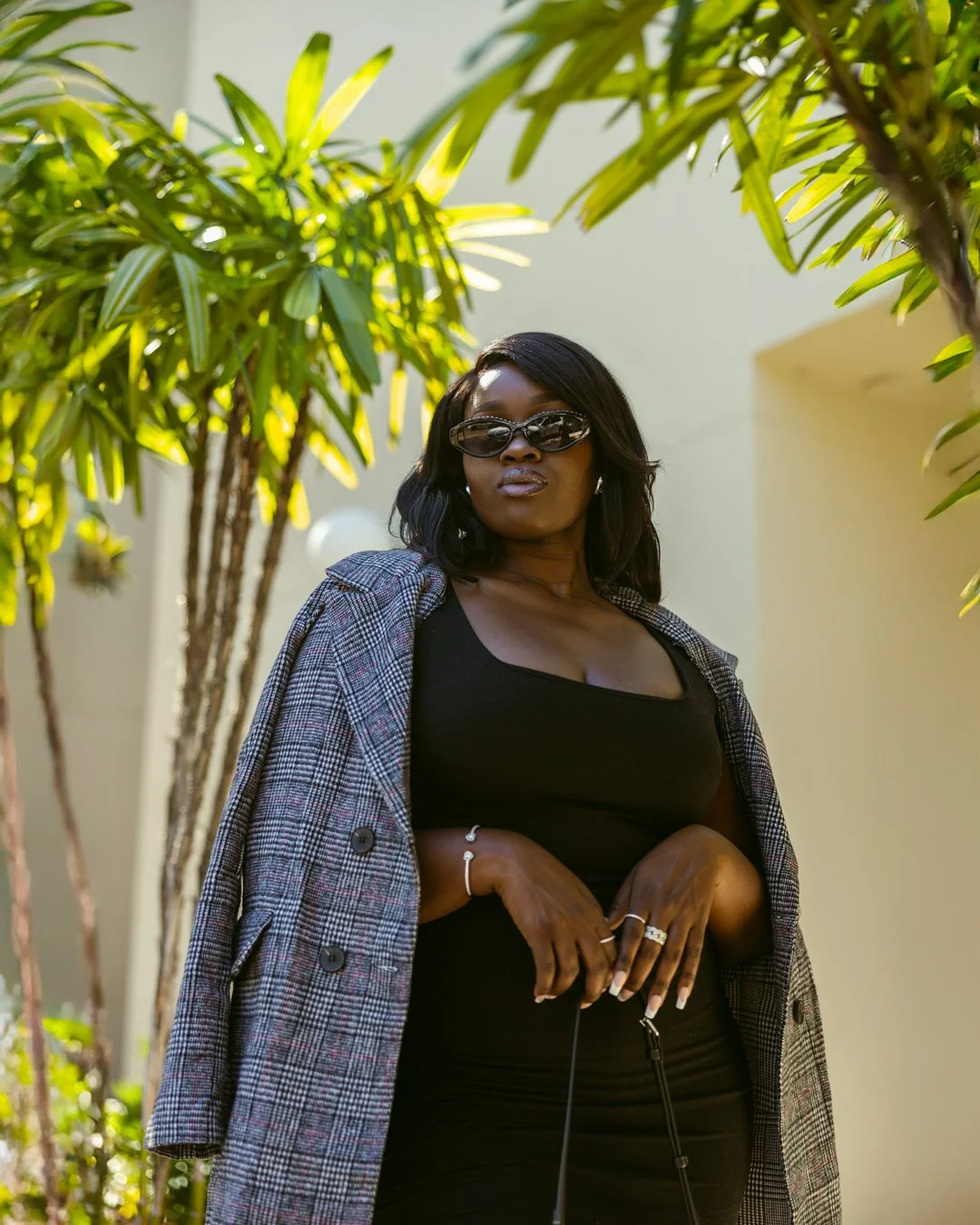 A confident woman wearing sunglasses, a black top, a plaid blazer, and jewelry stands outdoors amid green plants.