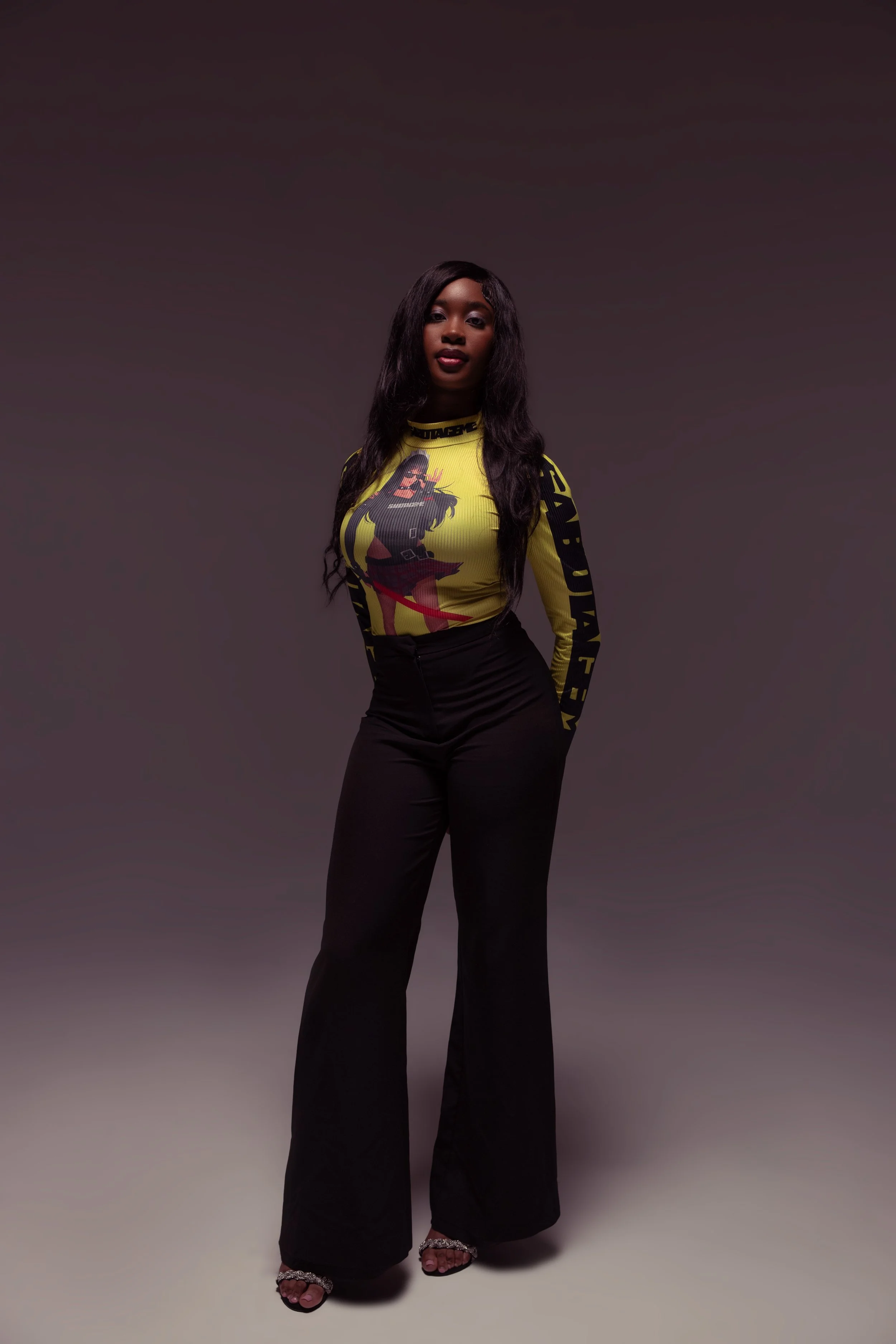 A woman with long dark hair stands against a gradient background wearing a yellow top with black text and graphics, black pants, and silver high-heeled sandals.