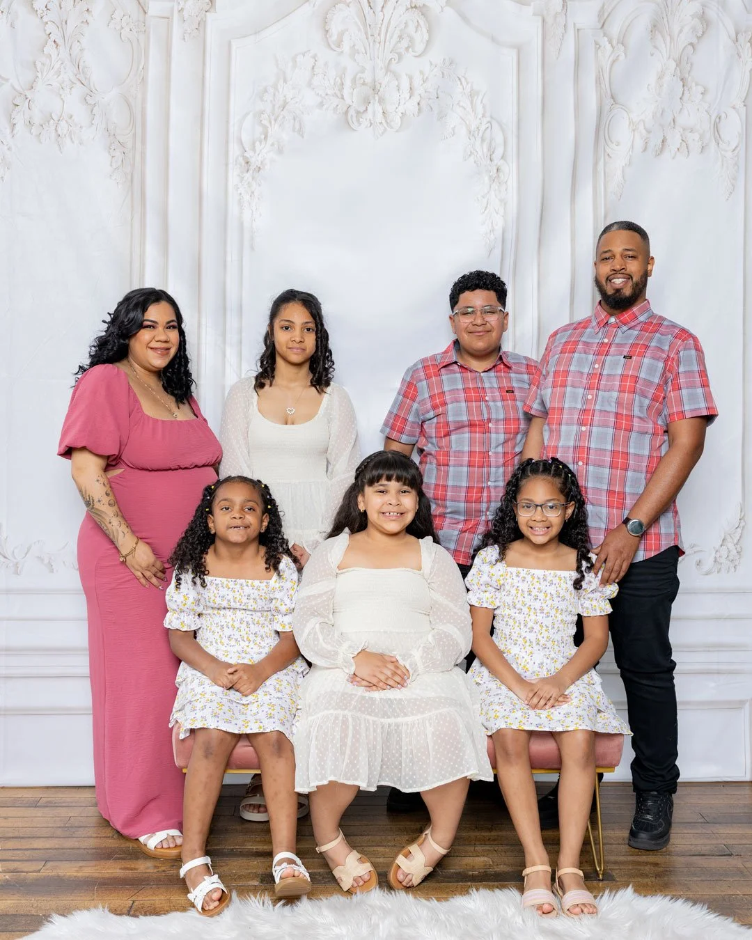 A large family portrait in front of a white ornate wall. Four children are seated on a pink bench, with three women and two men standing behind them. The family members are smiling and dressed in casual and semi-formal clothing.