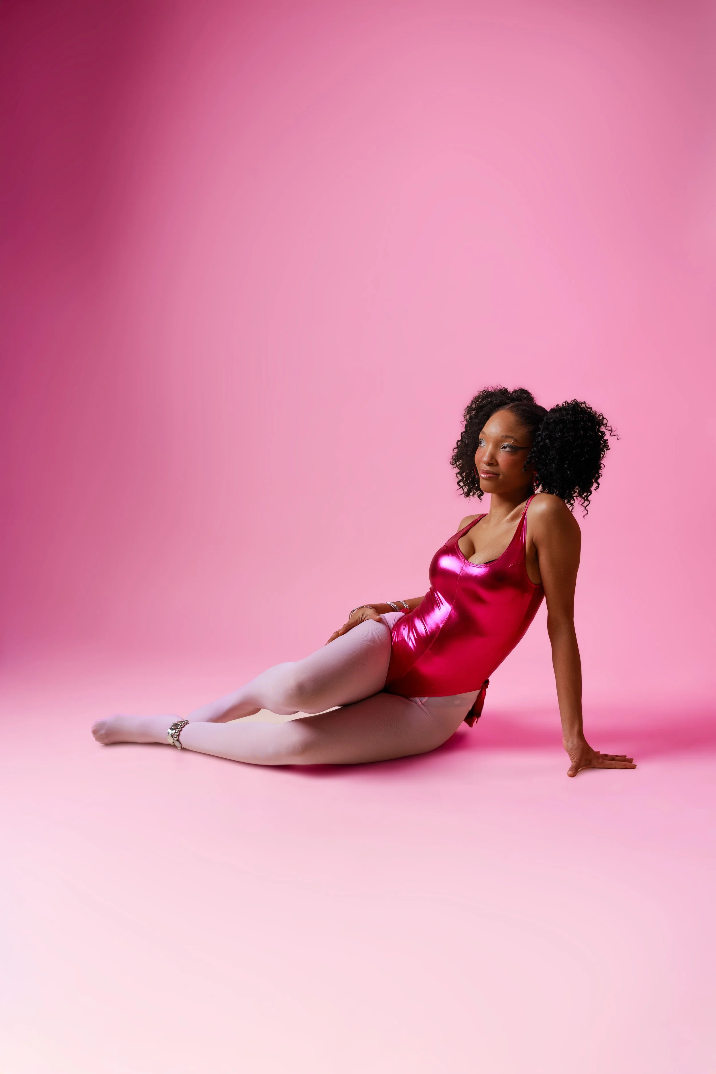 A woman with curly hair sitting on a pink floor against a pink background, wearing a shiny pink dress, white tights, and ballet slippers, gazing to the side.