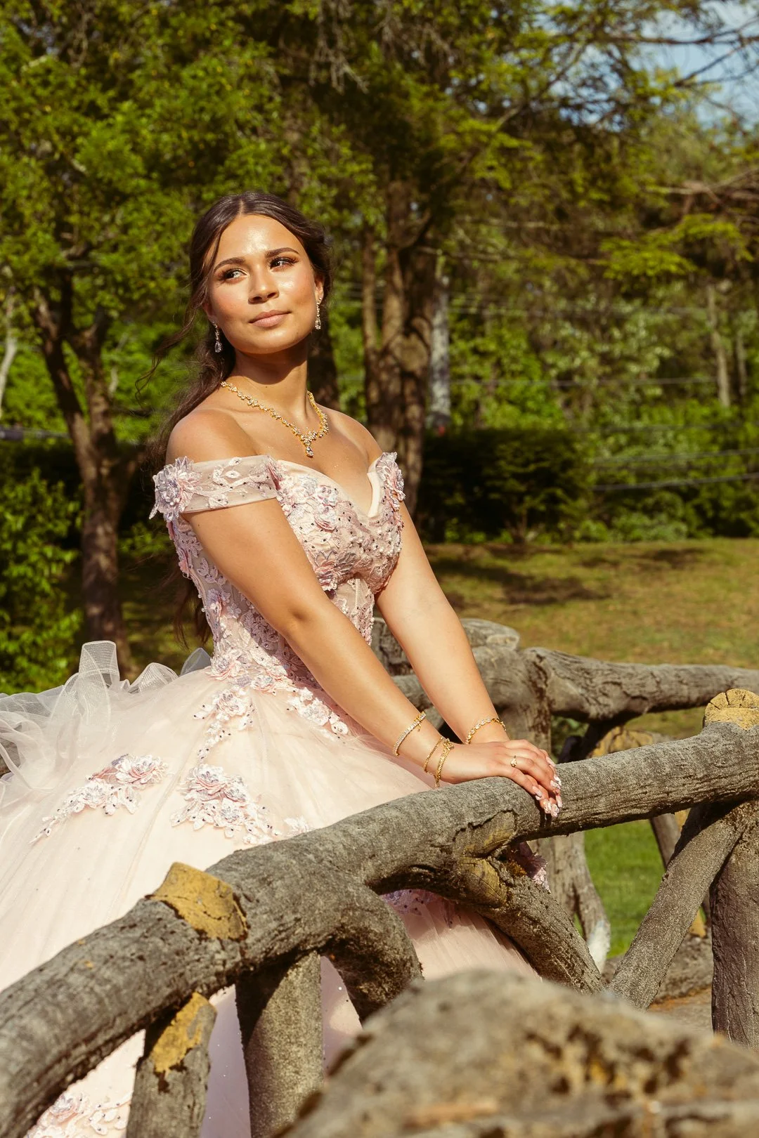 Young woman in a pink, floral, off-shoulder dress standing outdoors beside a wooden fence, with trees and greenery in the background.