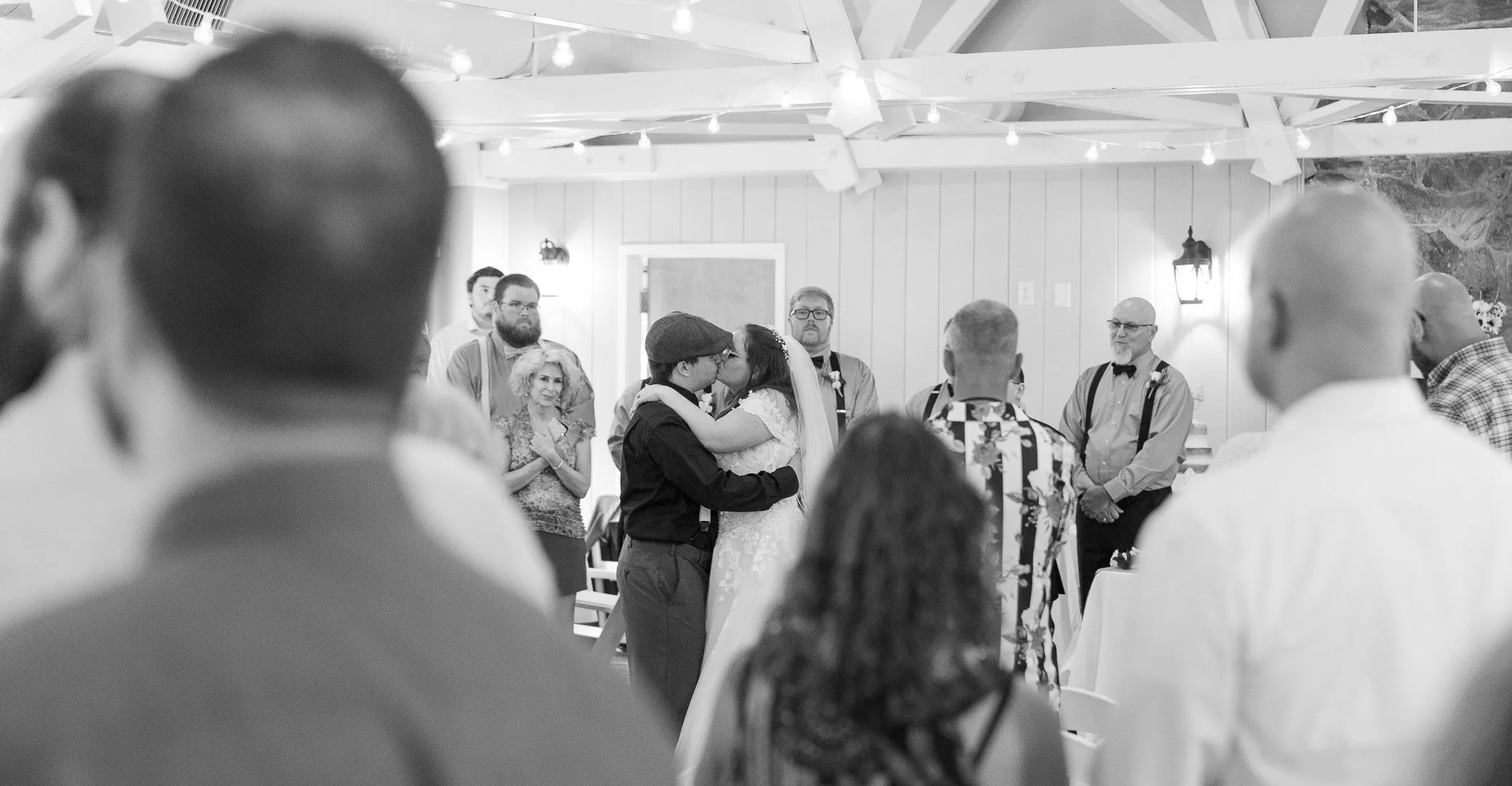 A black-and-white photo of a wedding ceremony where the bride and groom are sharing a kiss, surrounded by guests. The setting is indoors with string lights, a lantern, and rustic decor.