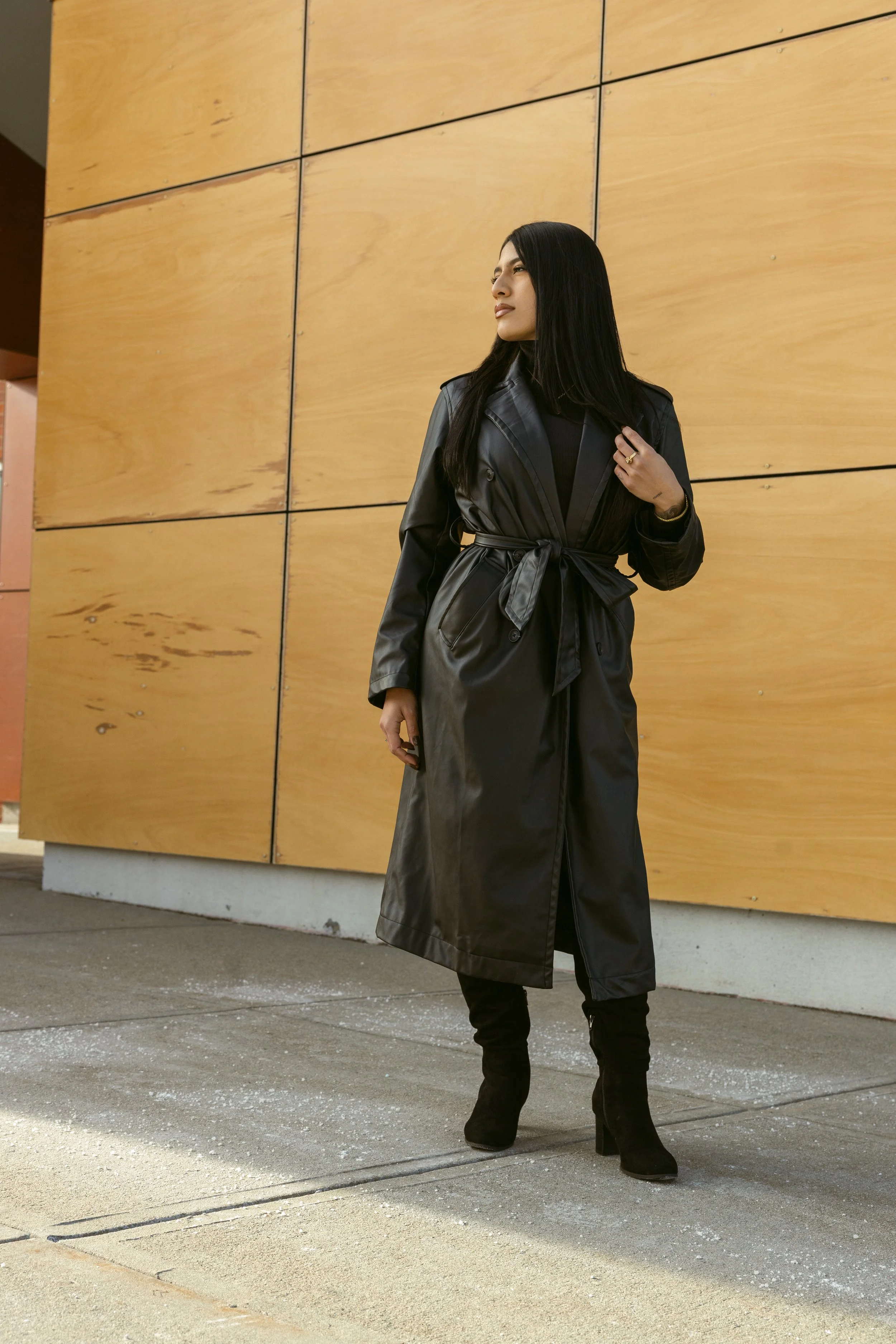 A woman with long black hair wearing a black leather trench coat, black heeled boots, standing outdoors against a wooden wall, holding a phone in her right hand and looking to her left.