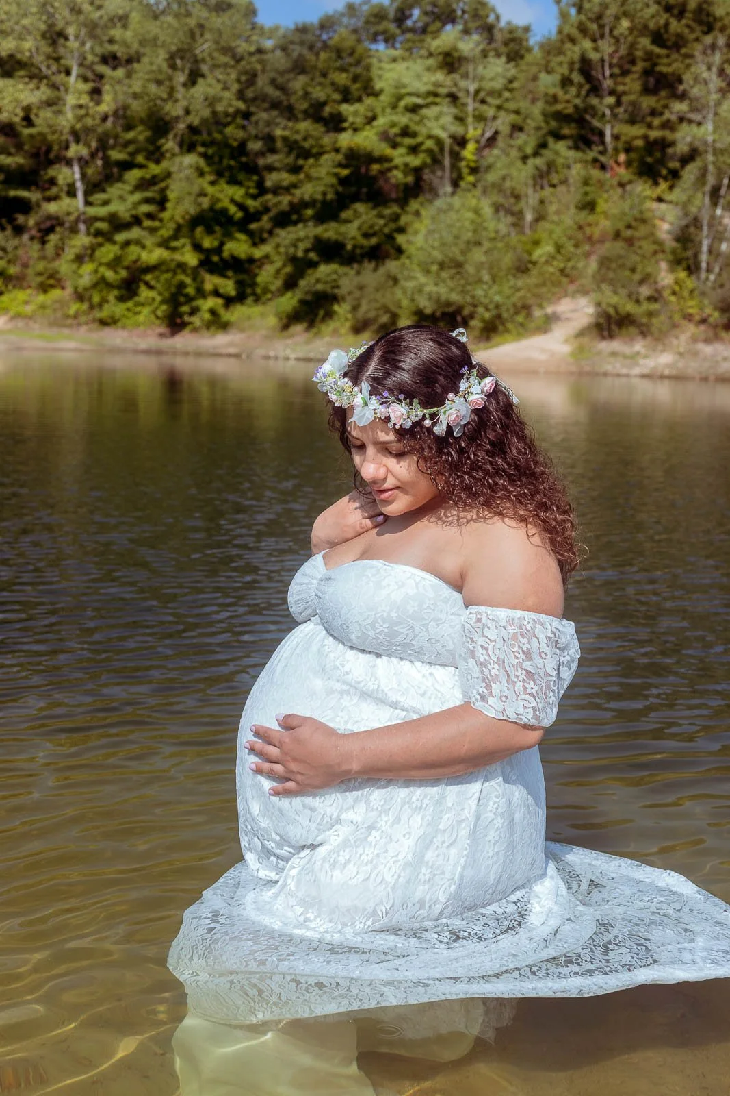 A pregnant woman wearing a white lace off-shoulder dress and a floral crown kneeling in a calm lake surrounded by green trees.