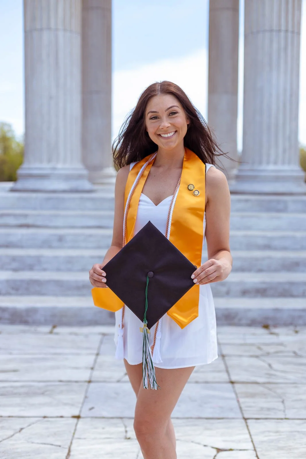 A smiling young woman in a white dress and a gold graduation stole holding a graduation cap standing in front of marble columns at a graduation ceremony.