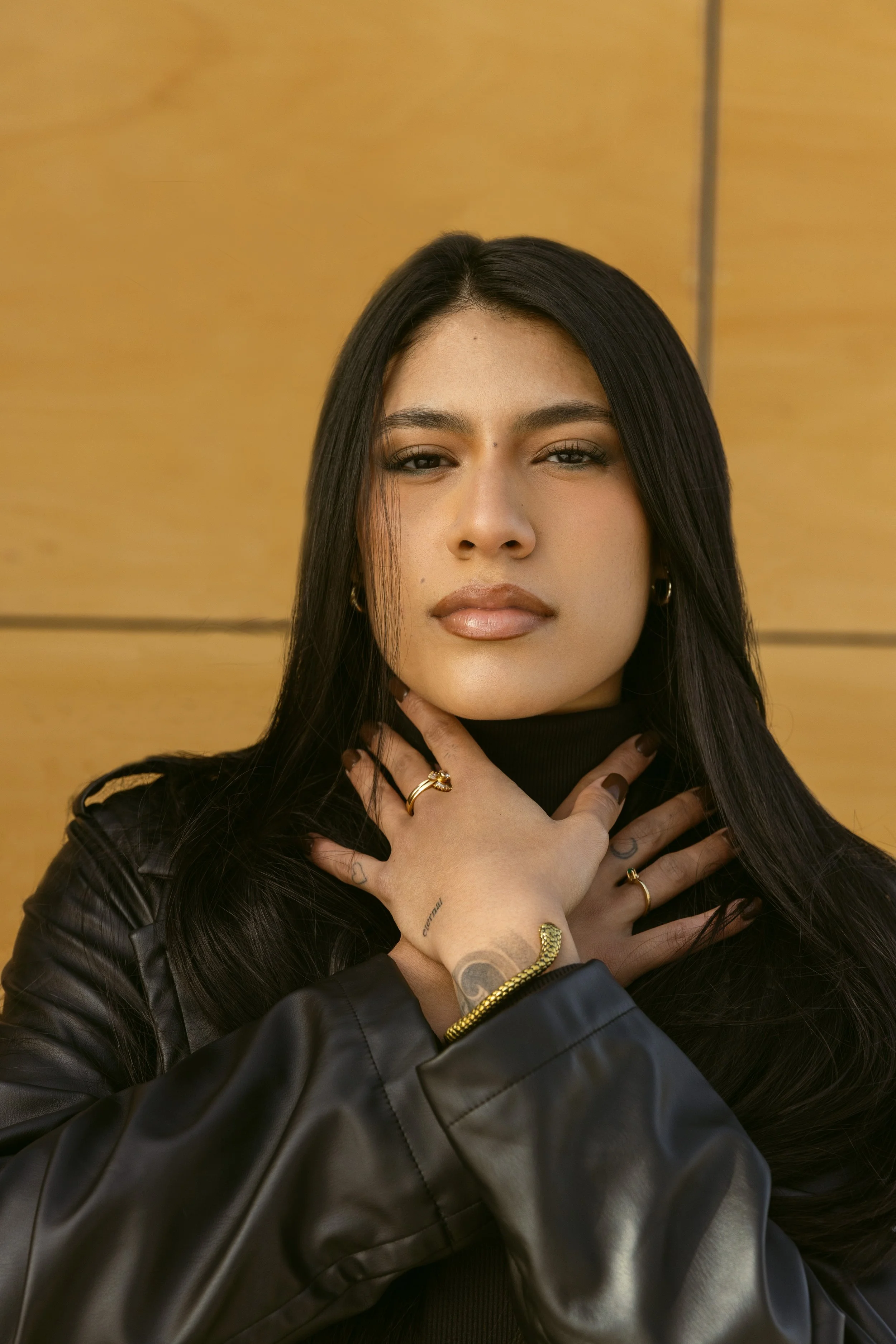 A young woman with long dark hair and tan skin poses with her hands crossed over her chest, wearing rings and a black leather jacket against a plain yellow background.