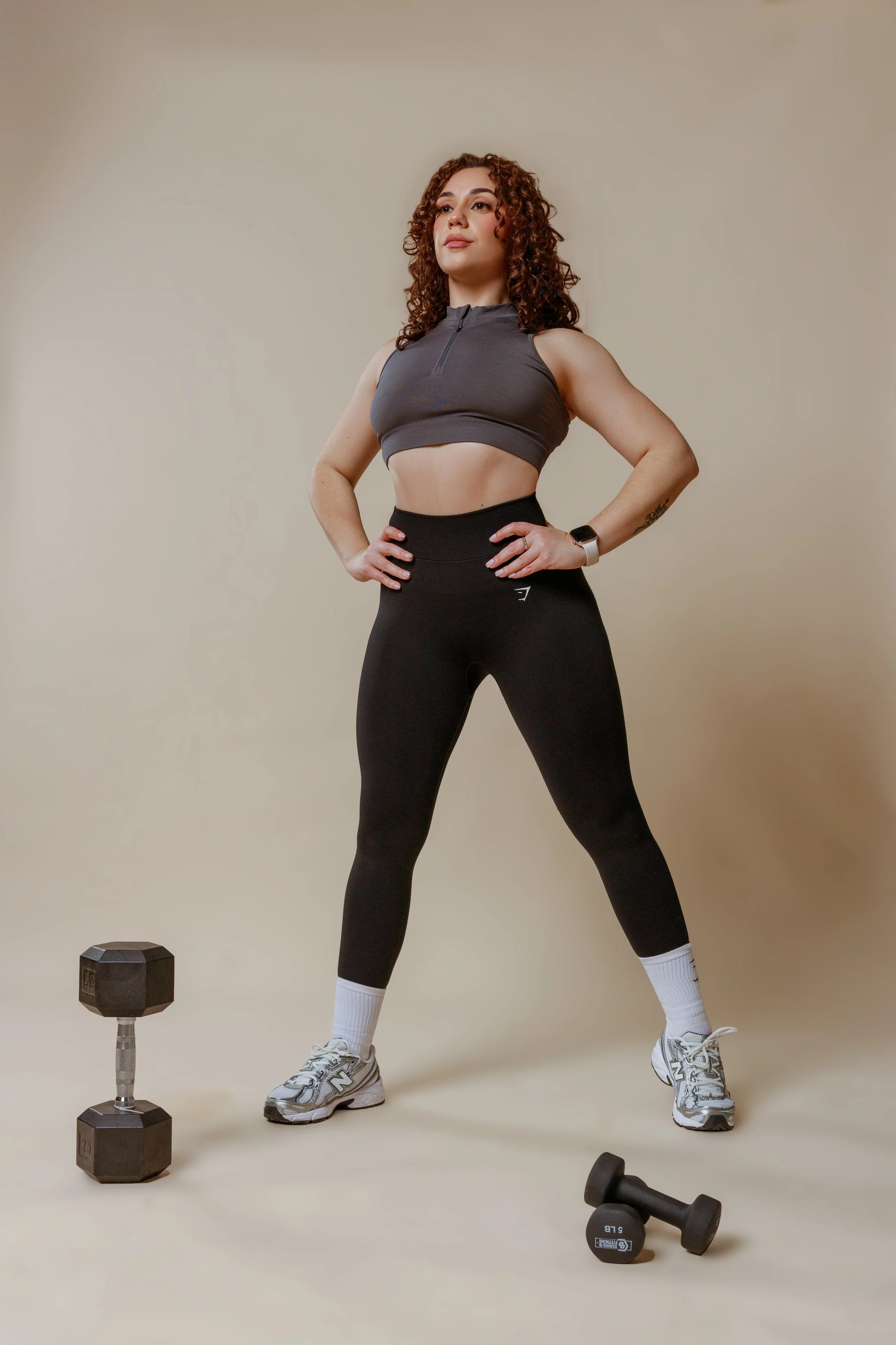 A woman with curly red hair, wearing a black athletic crop top and black leggings, stands with hands on hips in a workout setting. Dumbbells are on the ground, including a hexagonal dumbbell near her left knee and smaller dumbbells at her right. The 