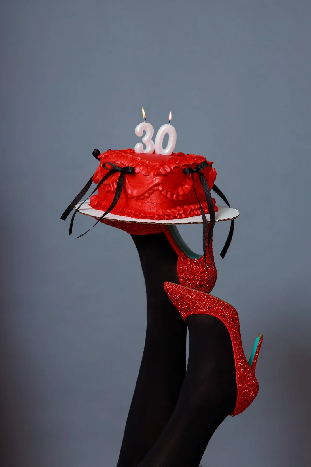 A person wearing red glitter high heels and black tights, holding a small, decorated red cake with a "30" candle on top, against a plain background.