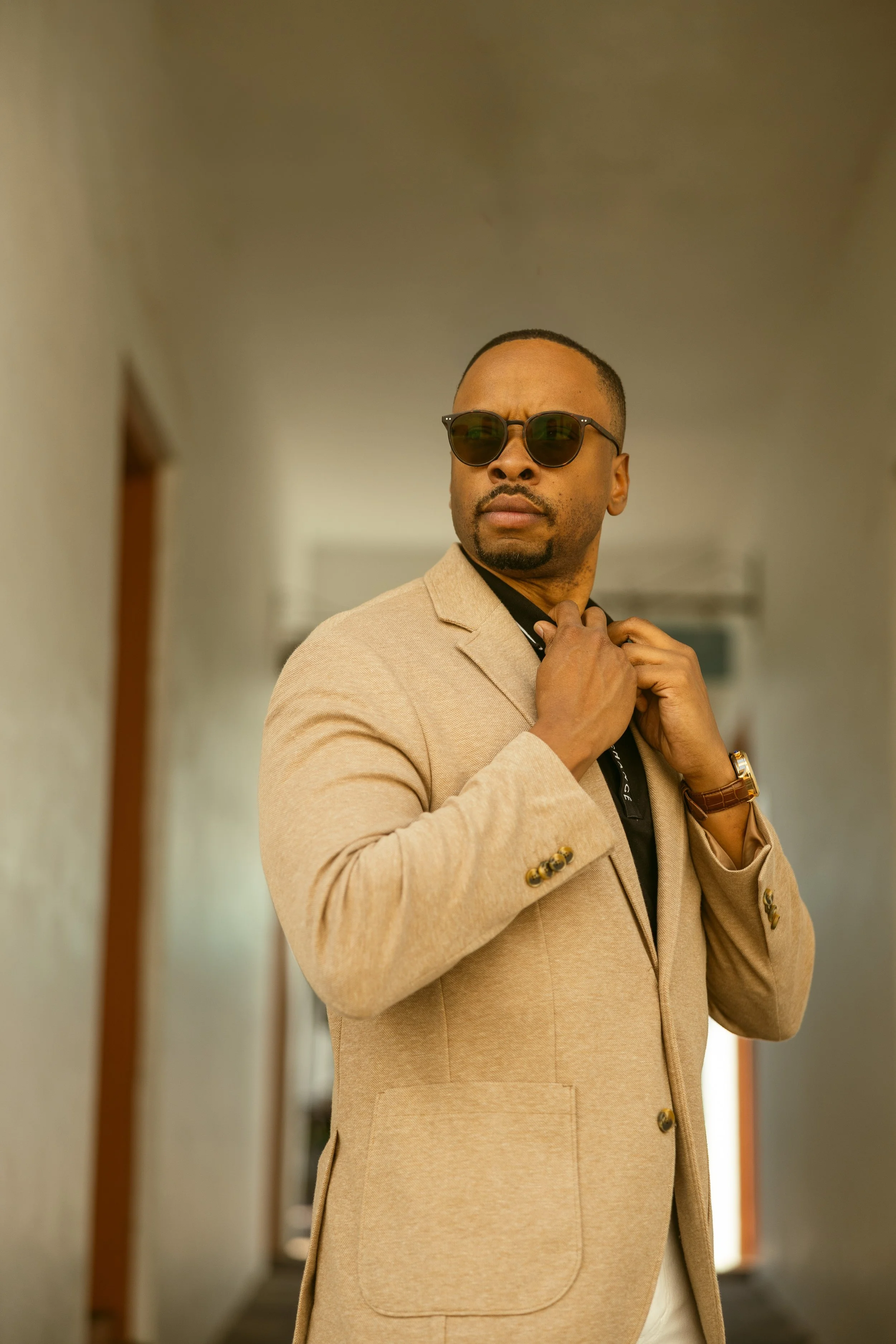 A stylish man adjusting his beige blazer in a hallway, wearing dark sunglasses, a watch, and a black shirt.
