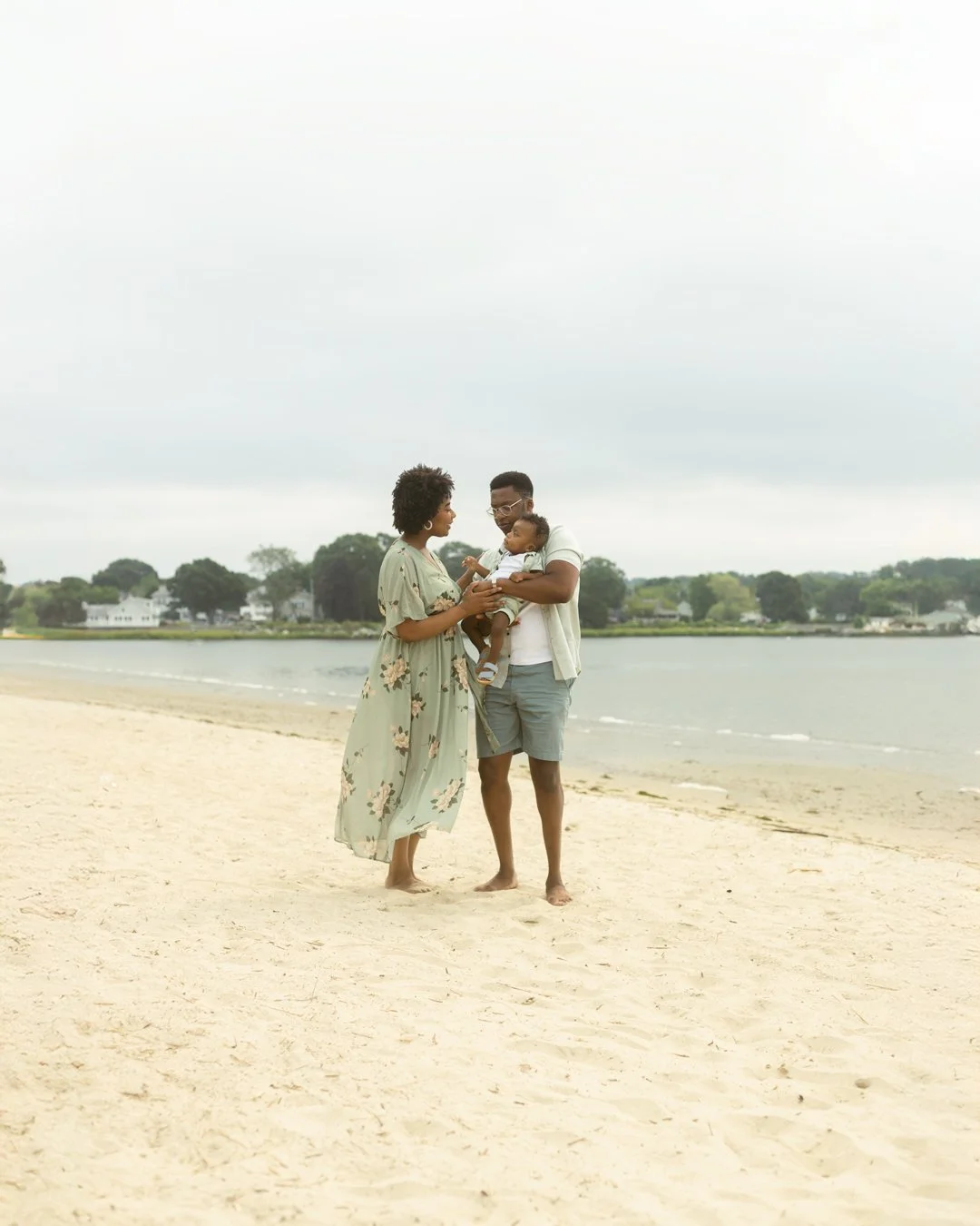 A family of three enjoying a moment together on a sandy beach, with the water and houses visible in the background.