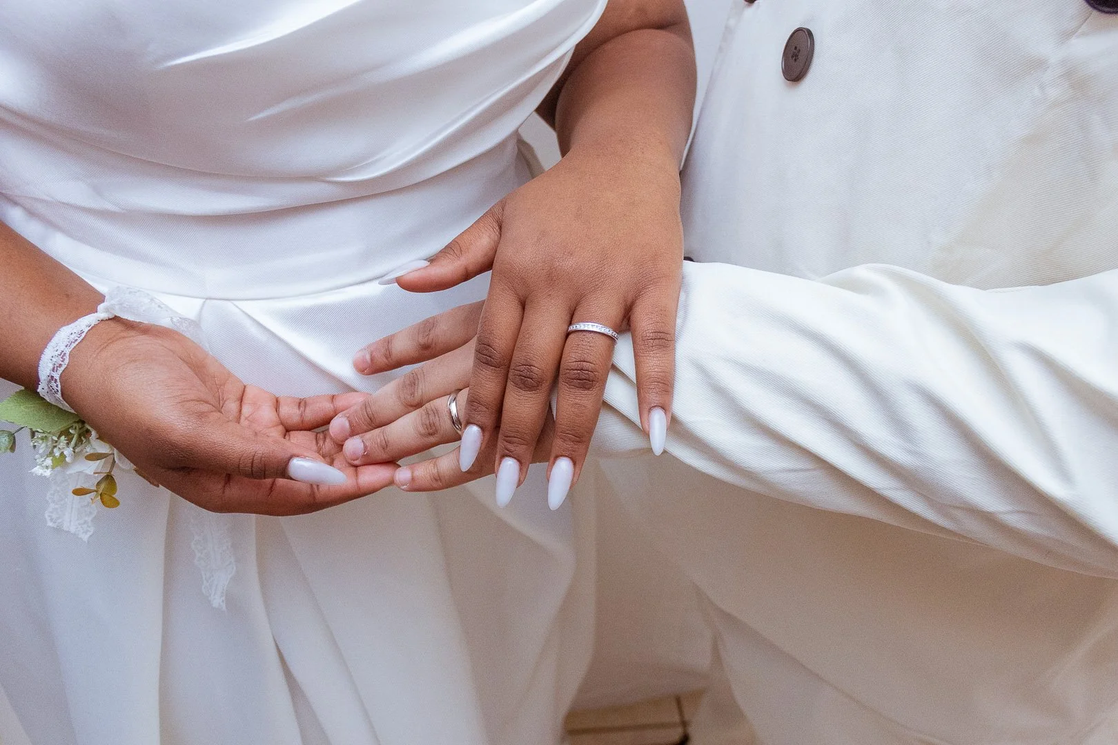 Close-up of two hands, one with a wedding ring, resting on the sleeve of a white wedding dress and a white suit, symbolizing a wedding or marriage ceremony.